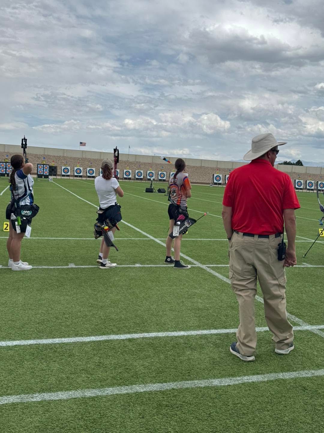A group of athletes stands on a lacrosse field near a coach in a red shirt under a cloudy sky.