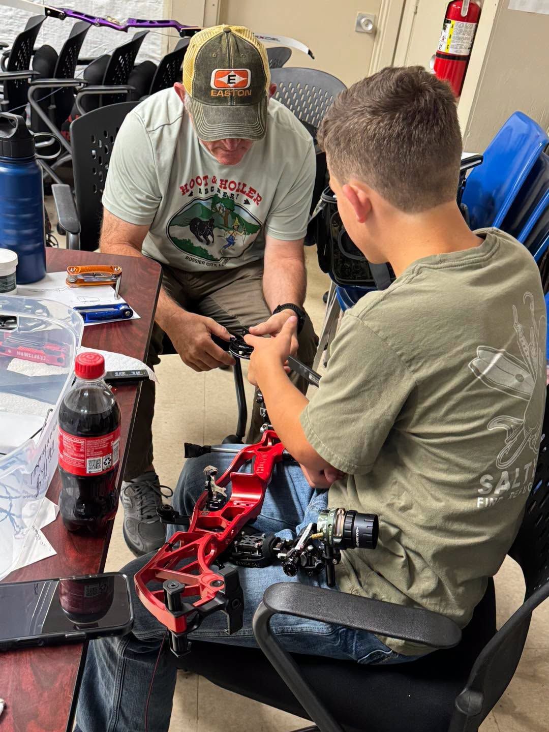 Two people work on a red mechanical assembly at a table with tools and soda bottles nearby