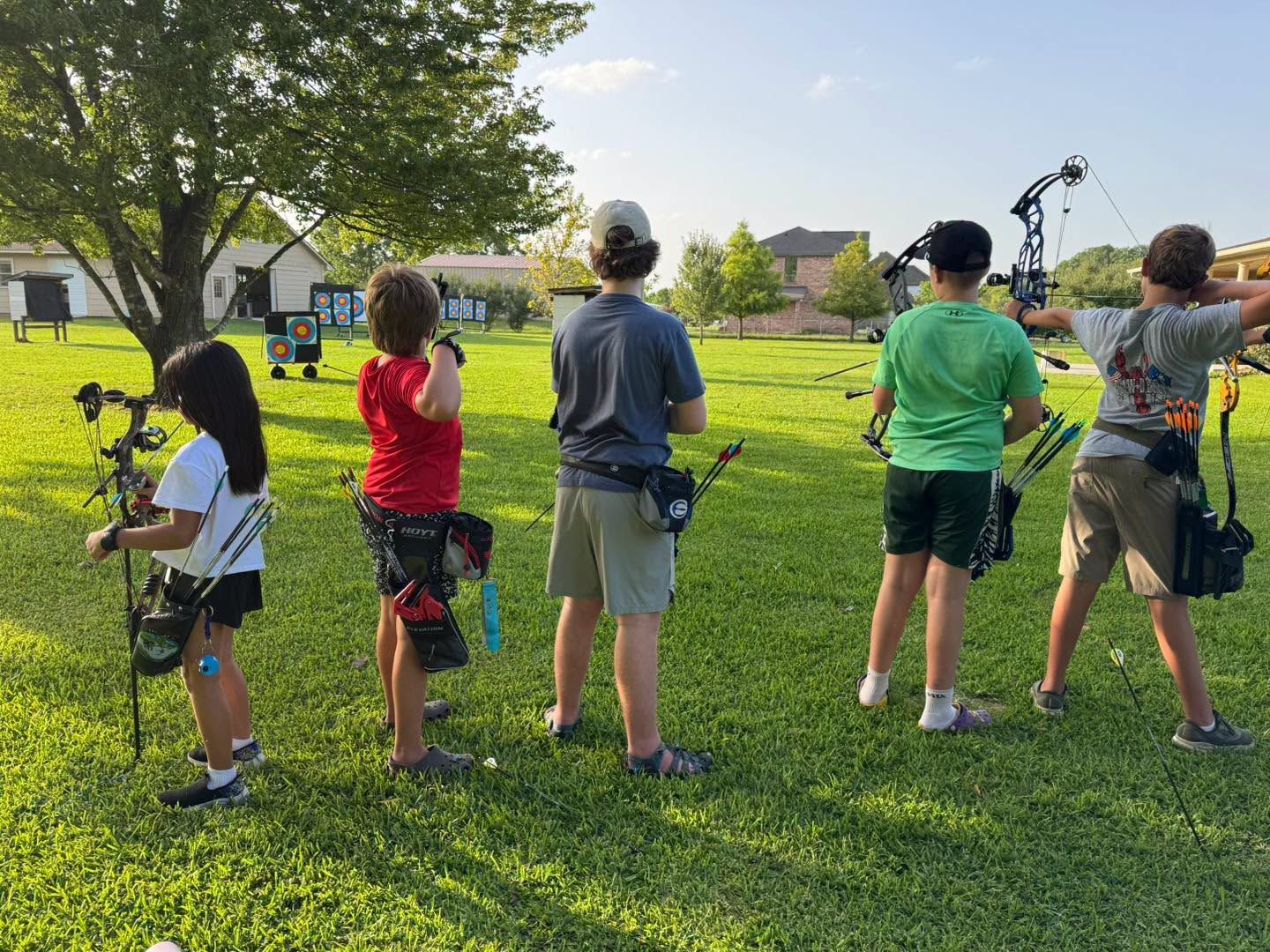 Children in a park practicing archery on a grassy field, backs to camera, with trees and houses in the background