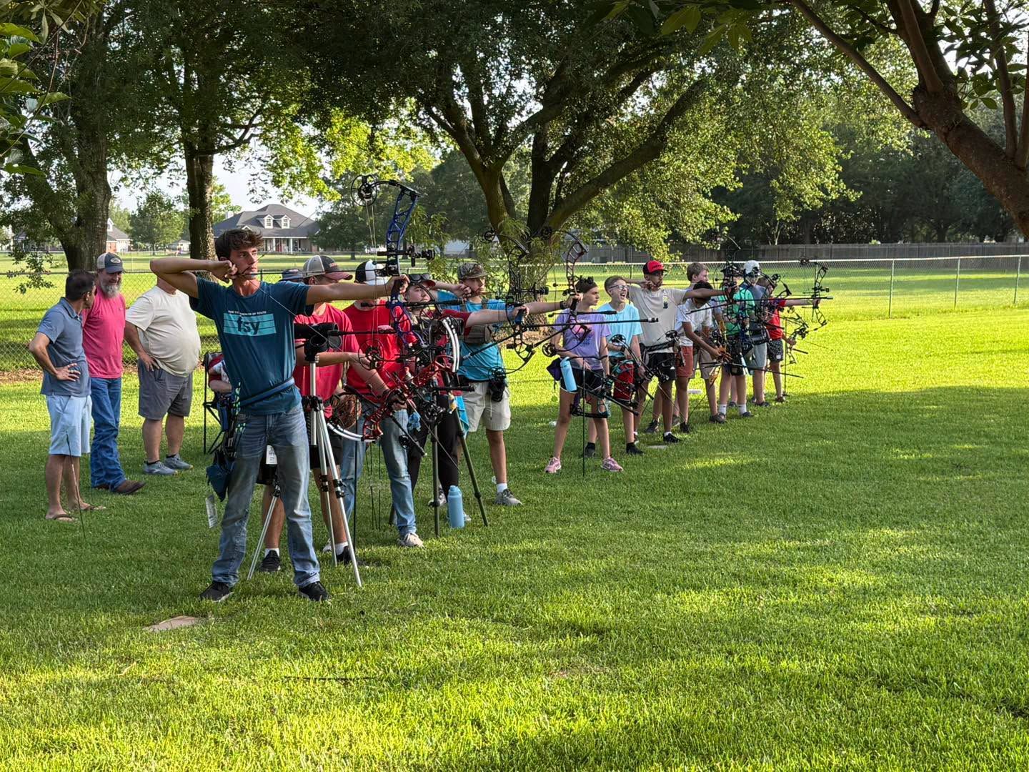 Children riding bicycles in a park, lined up on grass under trees with adults watching nearby