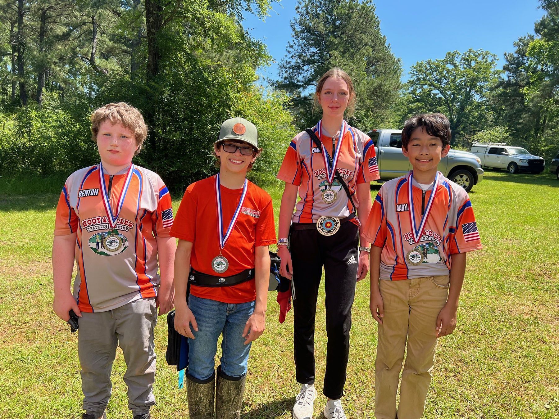 Four medal-wearing youths pose outdoors on grass under a sunny blue sky.