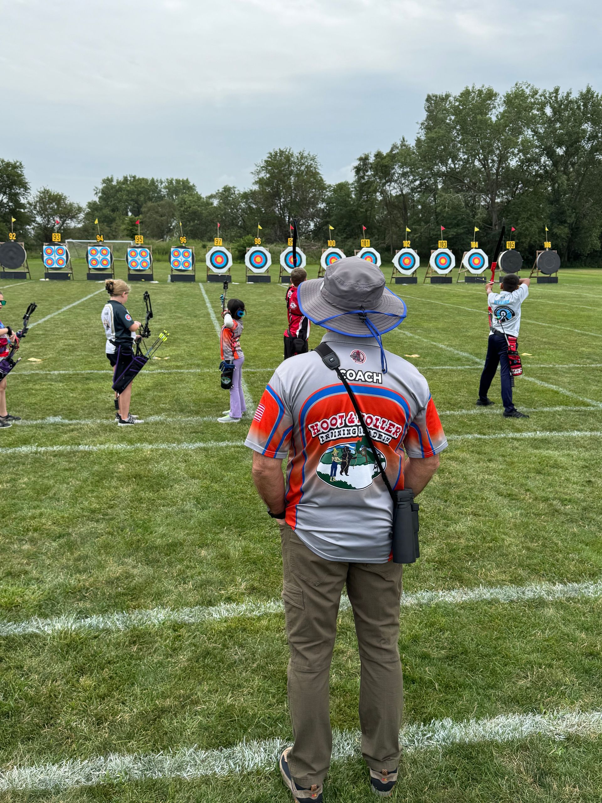 Archery practice on a grassy field, with archers lined up facing targets and an instructor in the foreground.