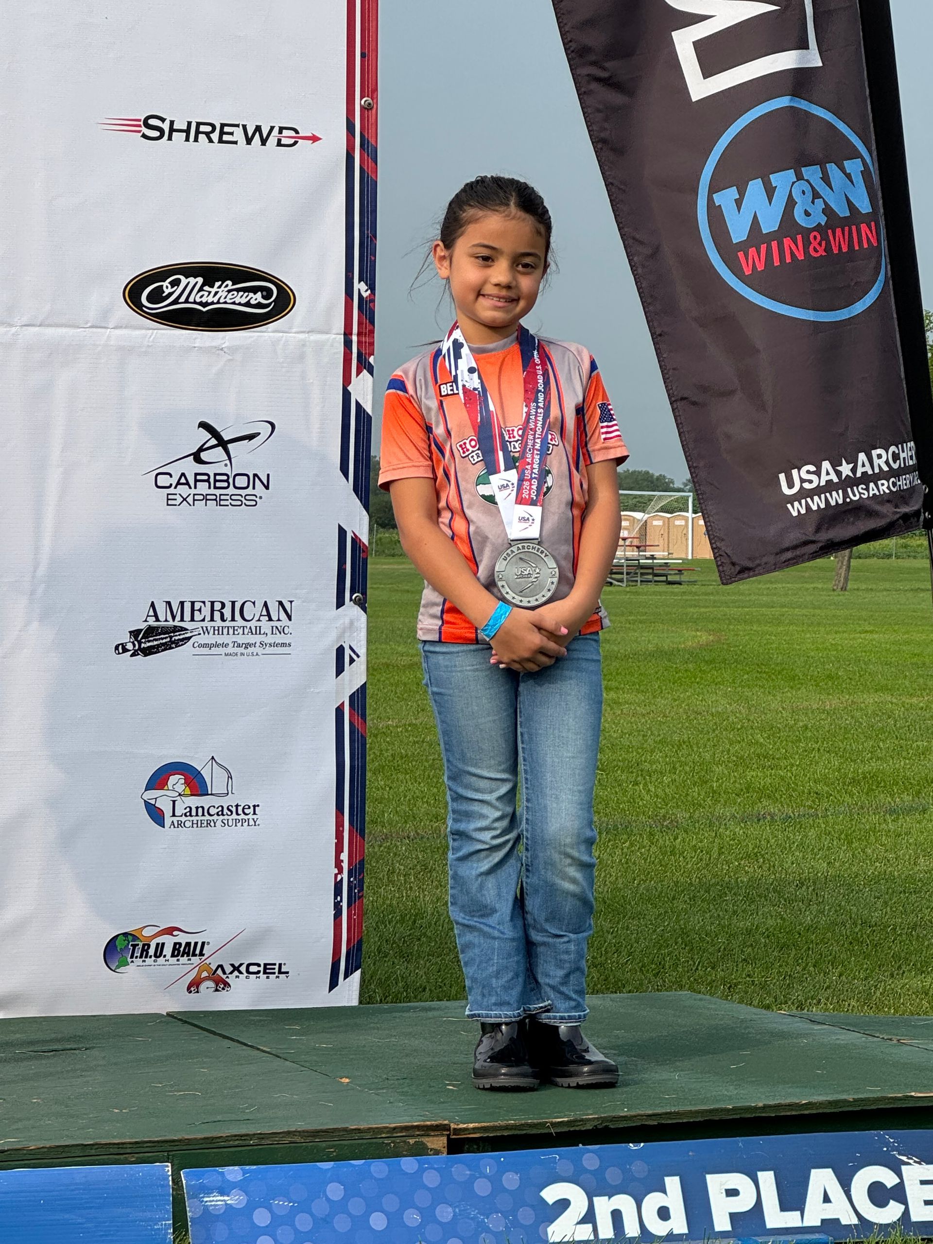 Child standing on a 2nd-place podium holding medals at an outdoor sports event