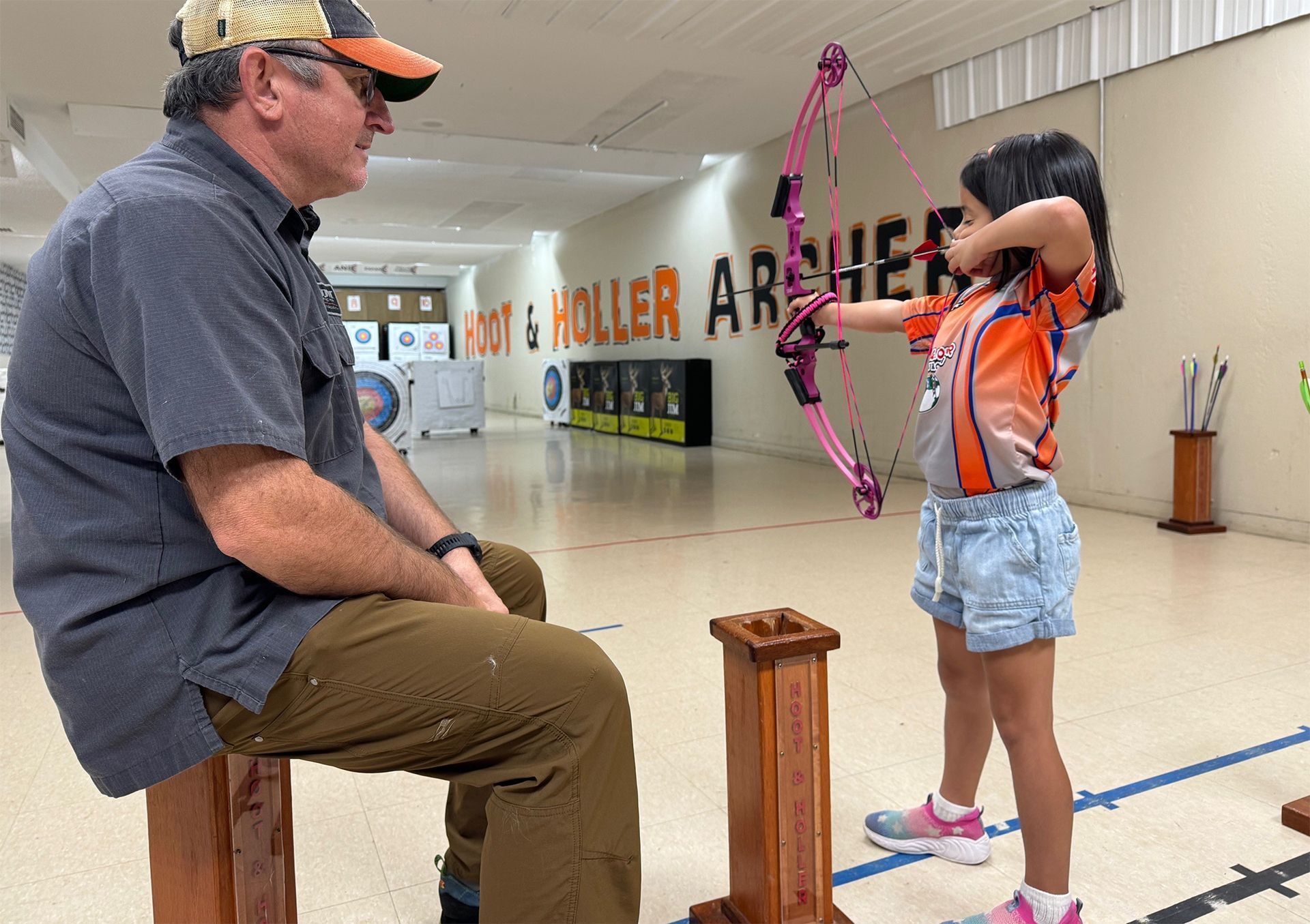 Man in cap seated indoors watches child practicing with a pink bow and arrow in a hallway