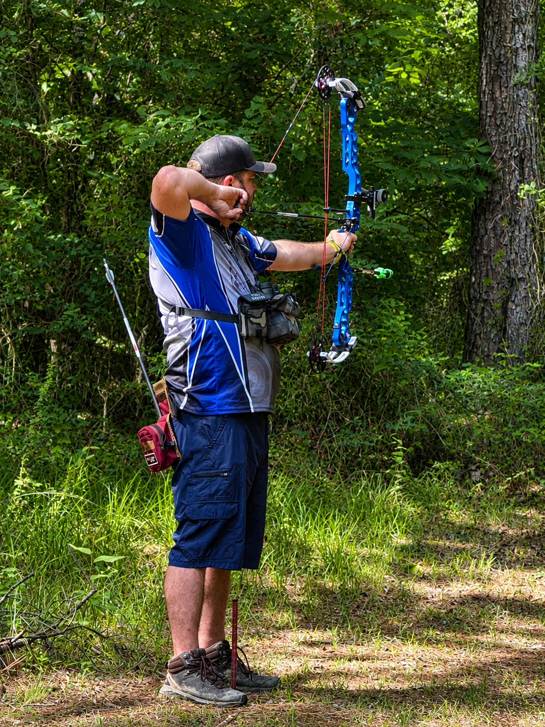 Person aiming a blue bow and arrow in a wooded area, wearing a blue shirt and dark shorts.
