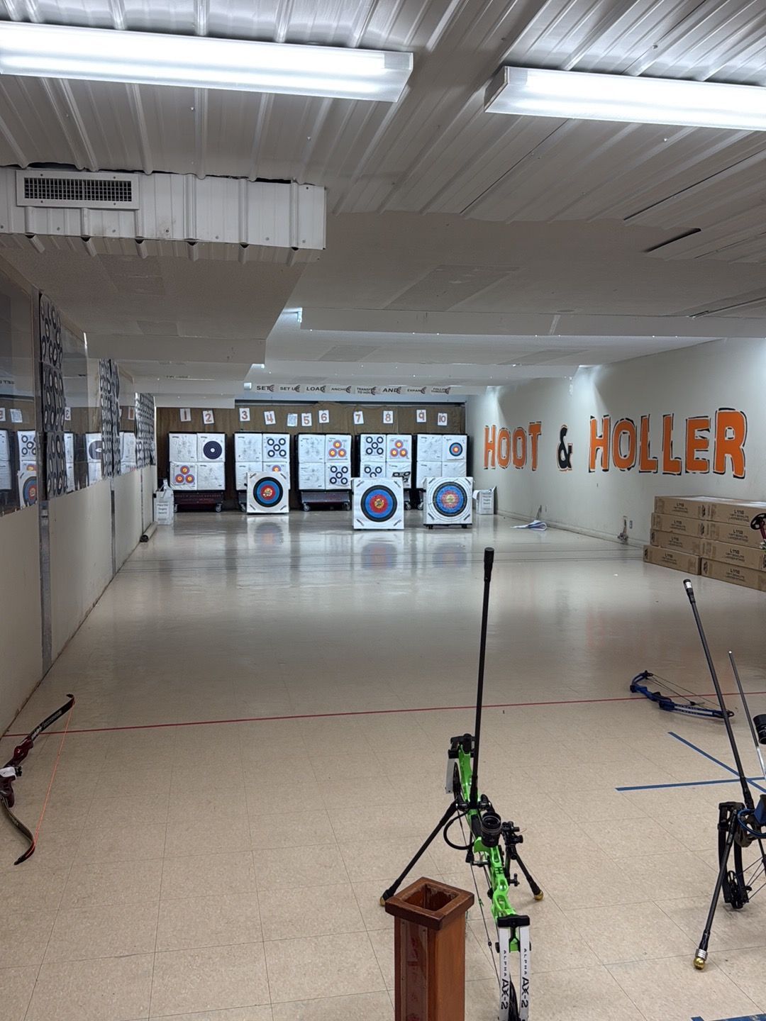 Indoor archery range with targets, bows, and shooting equipment laid out on a long lane.
