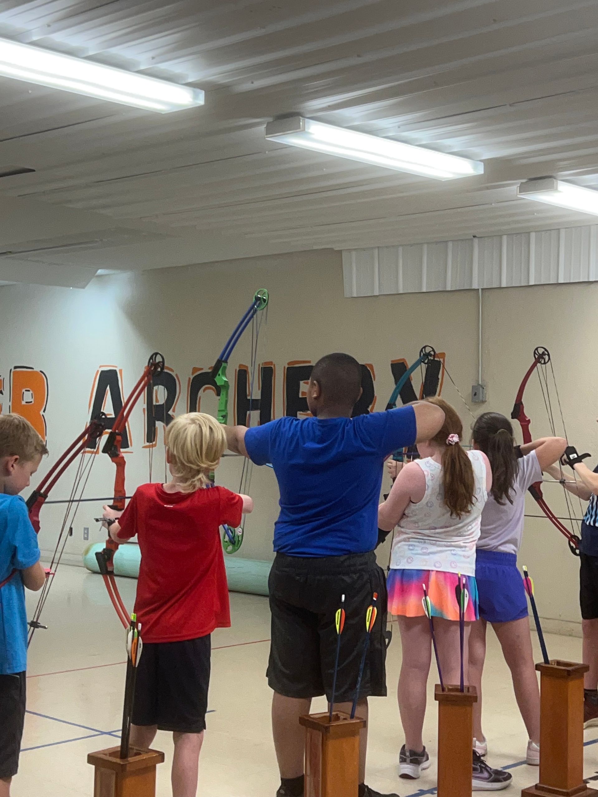 Children doing archery practice indoors, standing at targets with bows raised.