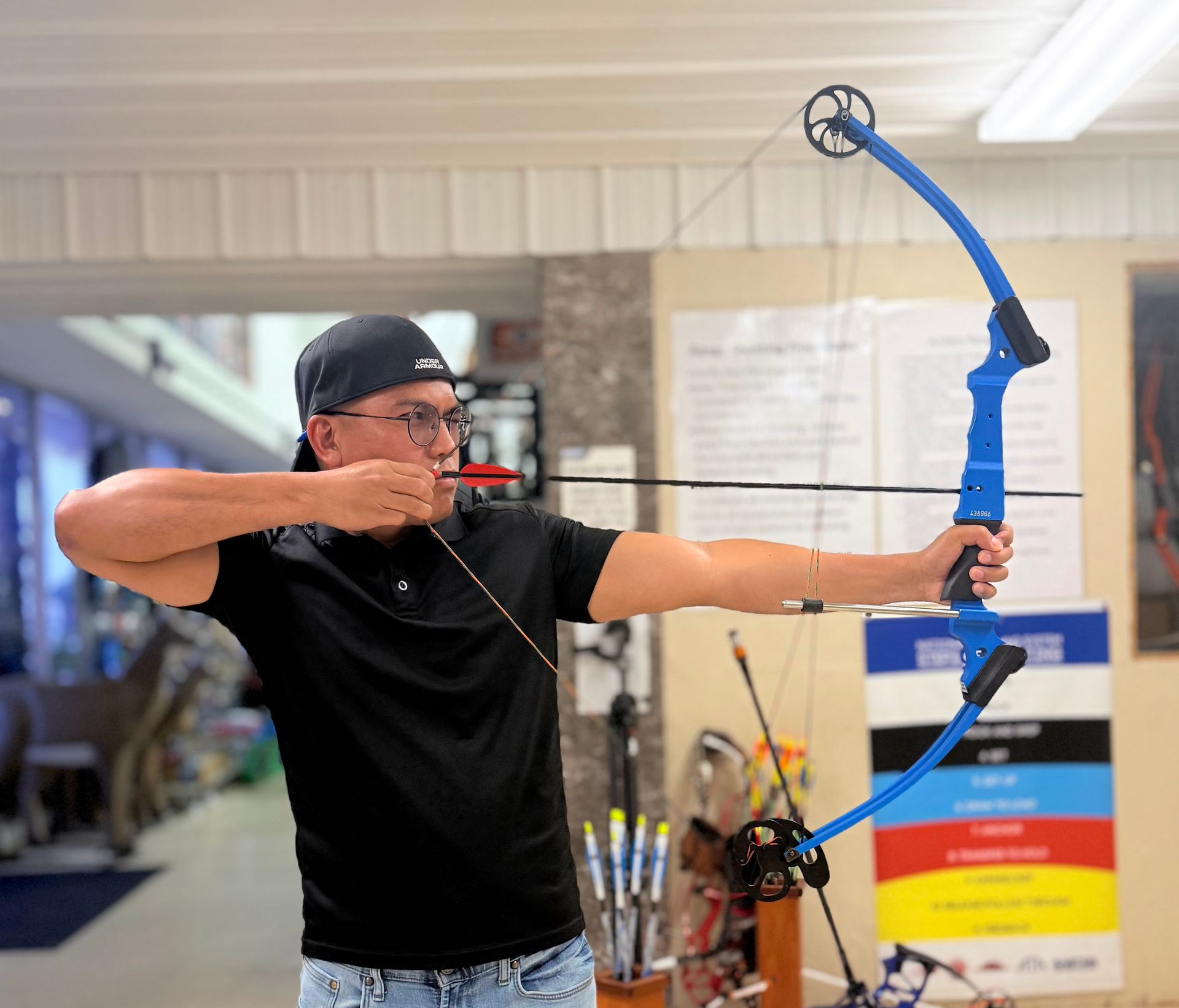 Person aiming a blue compound bow indoors, drawing the string in an archery range.