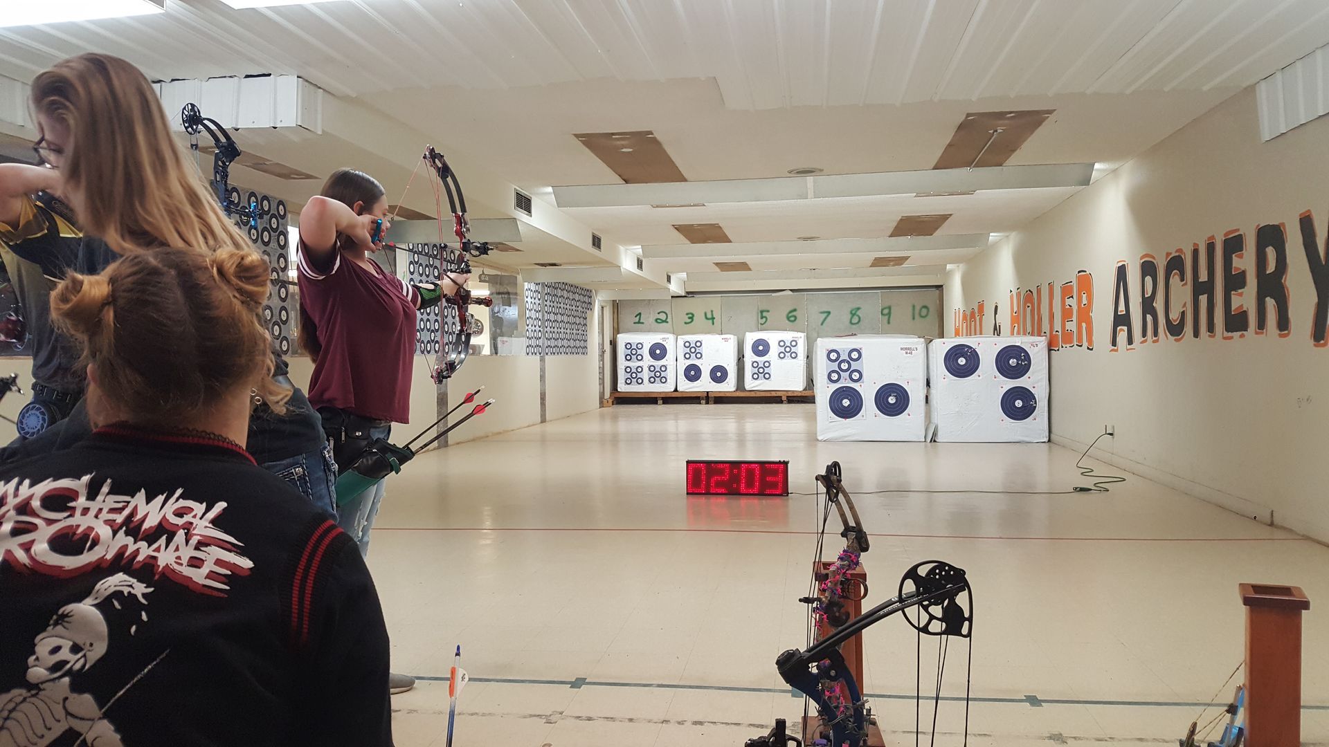 Archery range with several people aiming bows at distant targets in a bright indoor hall