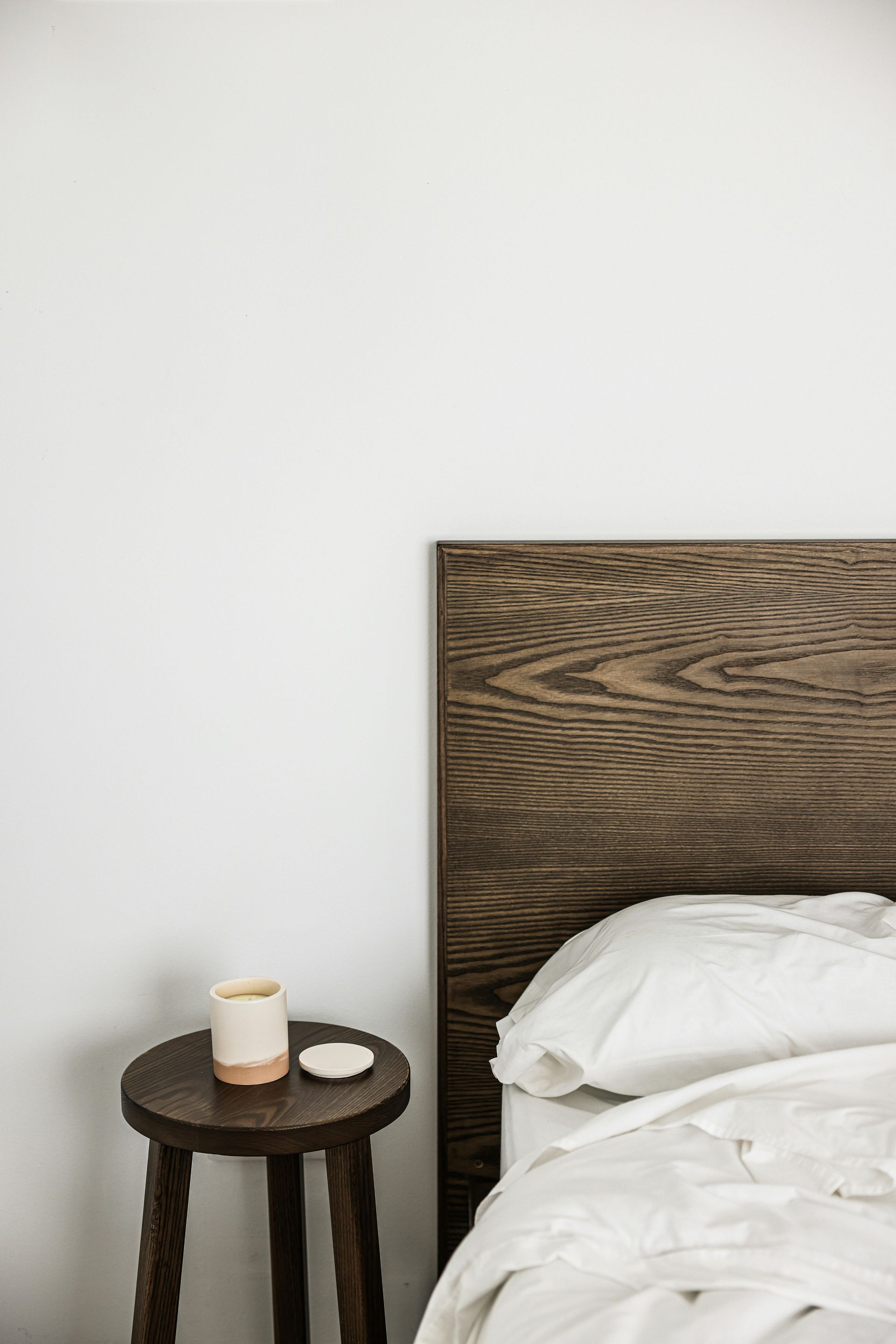 Wooden bed headboard and stool beside a bed with white linens, coffee cup.