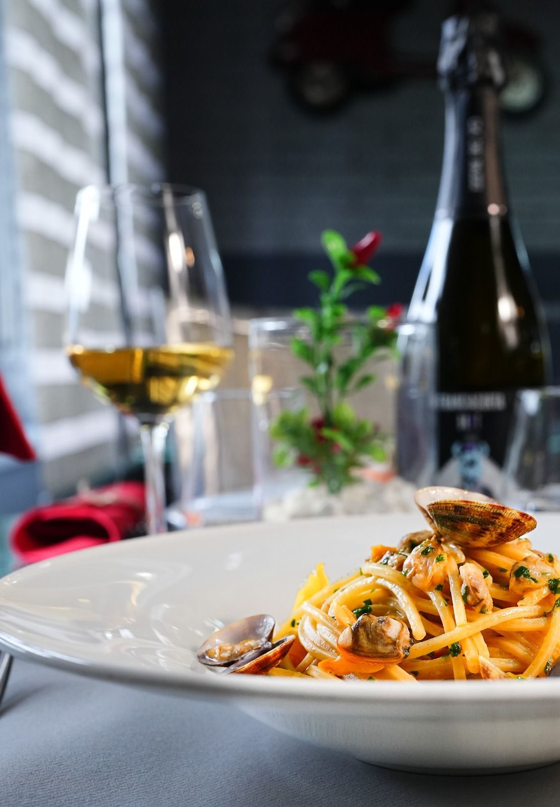 Piatto di pasta con le vongole, bicchiere di vino e bottiglia di spumante su un tavolo.