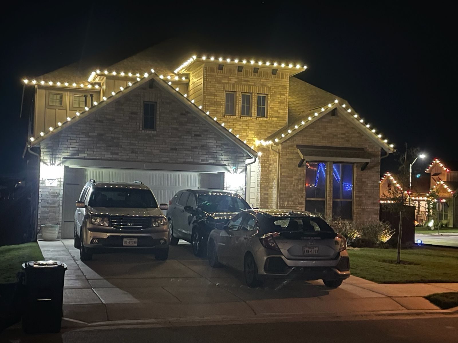 A house at night with Christmas lights on the roof; cars parked in the driveway.