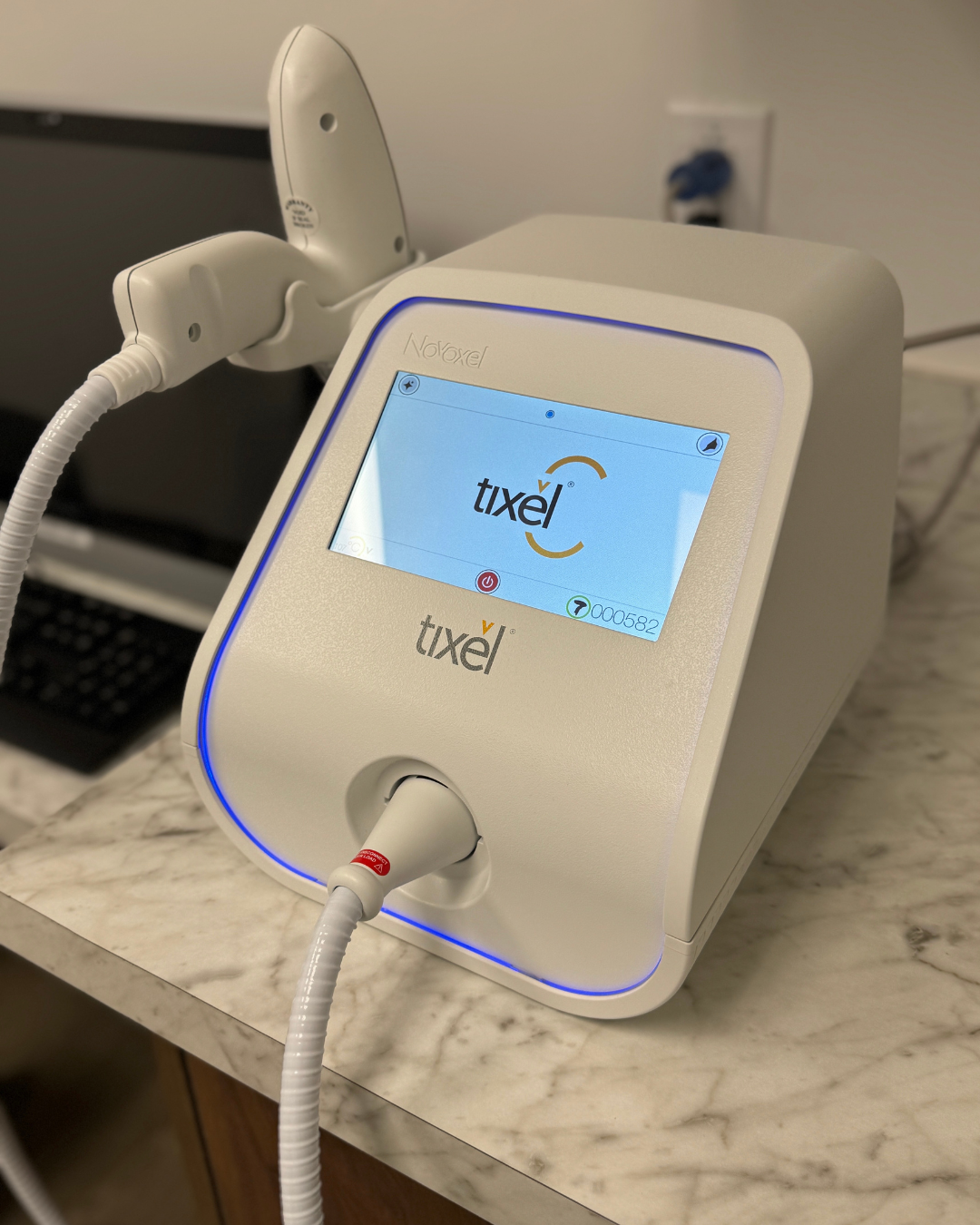 Tixel device, white with blue light, on a countertop. Attached handpieces and screen displaying the Tixel logo.