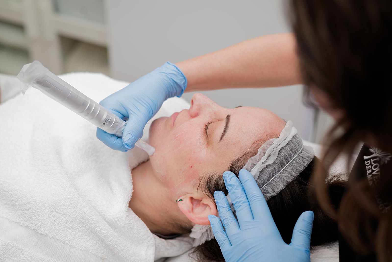 Woman receiving a facial treatment, held by gloved hands. Indoors, white cloth, light colors.