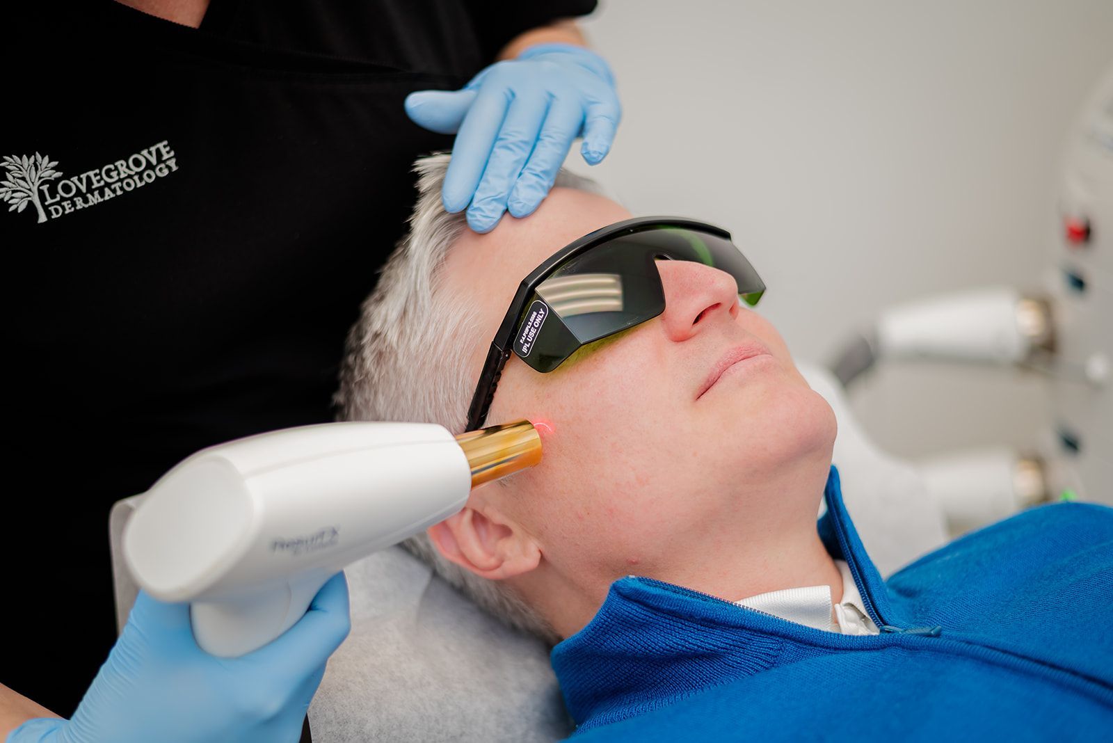 Man receiving laser facial treatment; practitioner in blue gloves holds device to his cheek.