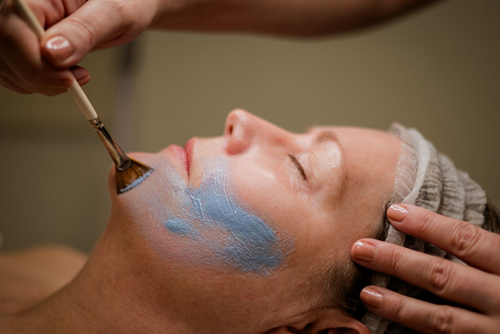 Woman receiving facial treatment, blue mask applied with a brush in spa setting.