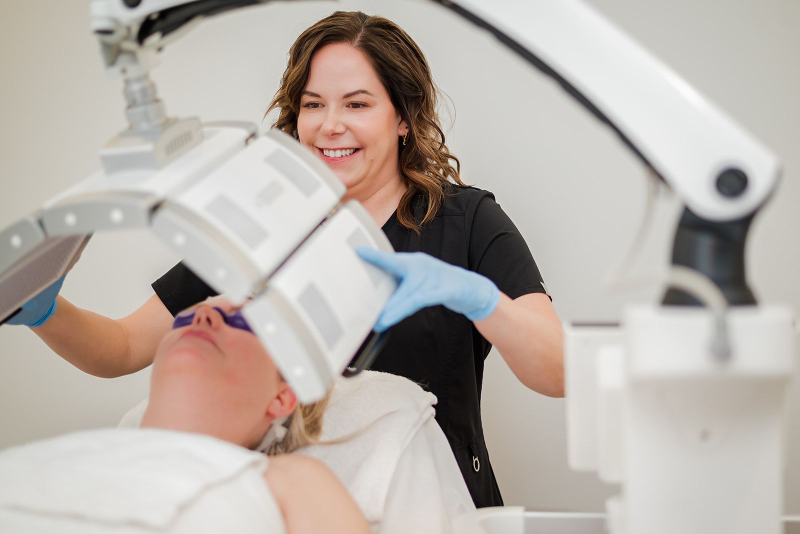 Aestehtician in a clinic administering blue light therapy to a patient. Both are smiling.