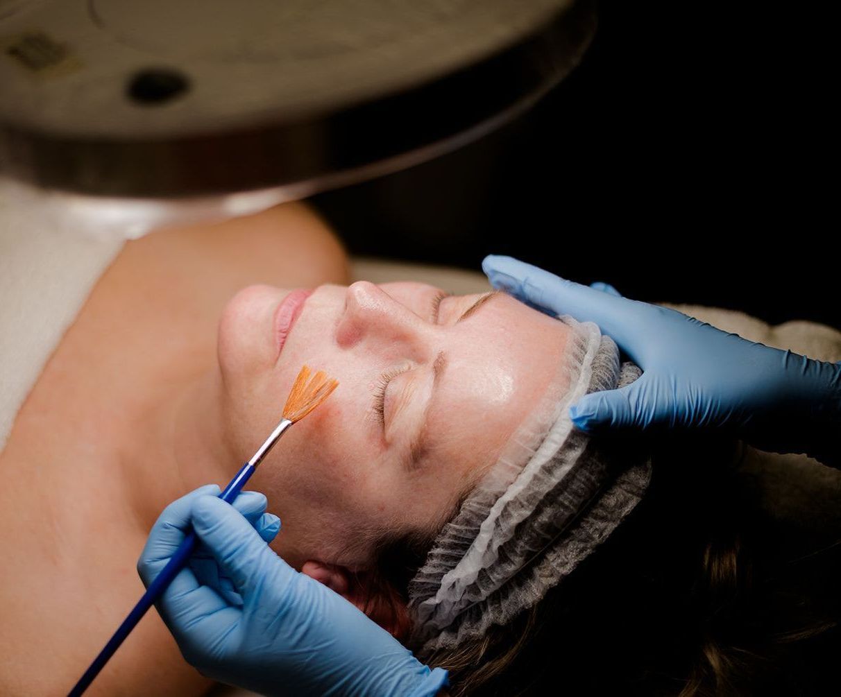 Woman receiving a facial chemical peel, being treated with a brush, under a magnifying lamp.