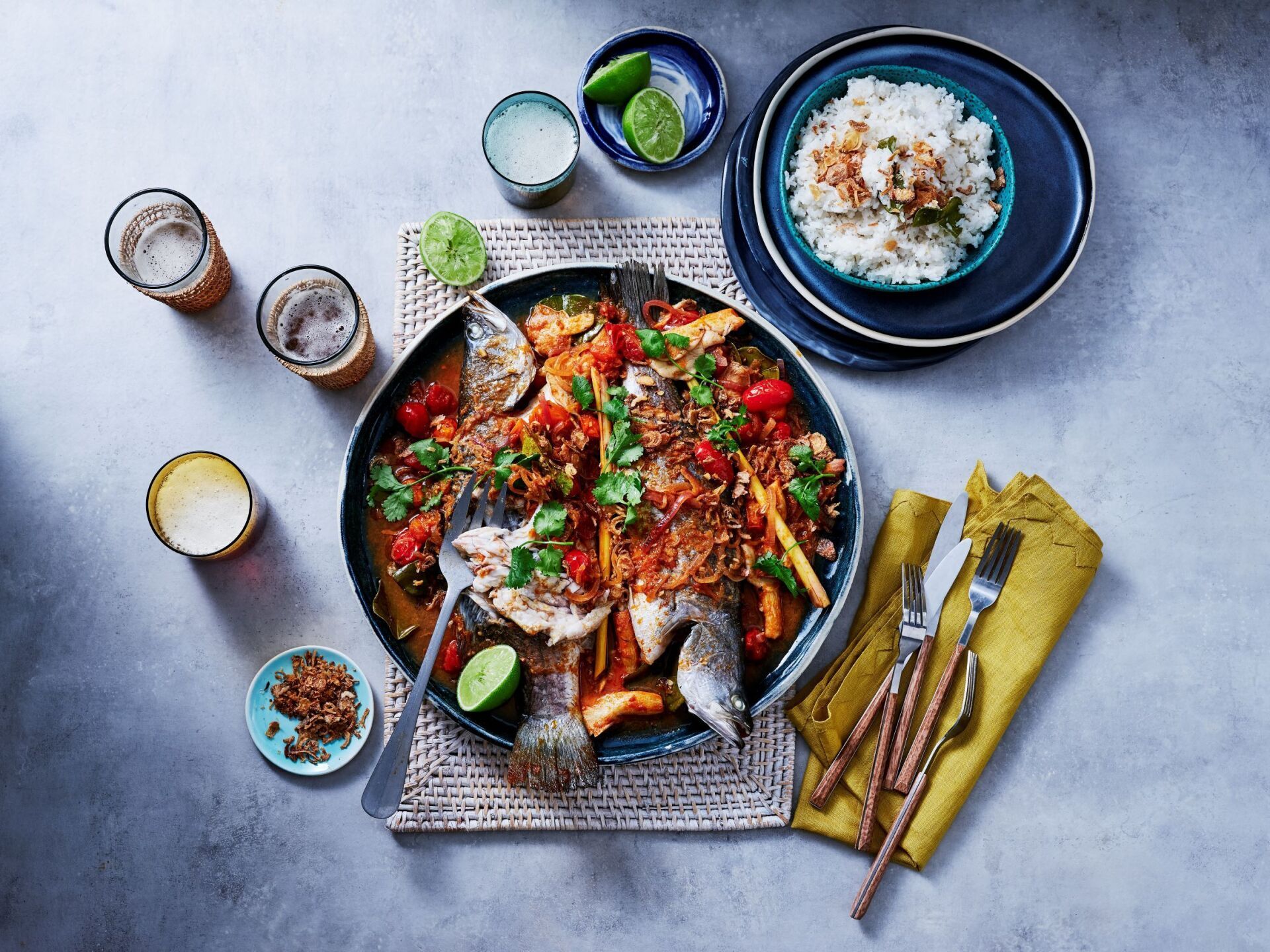 A large platter of cooked whole fish in a red sauce, served with rice, lime wedges, and glasses on a grey surface.