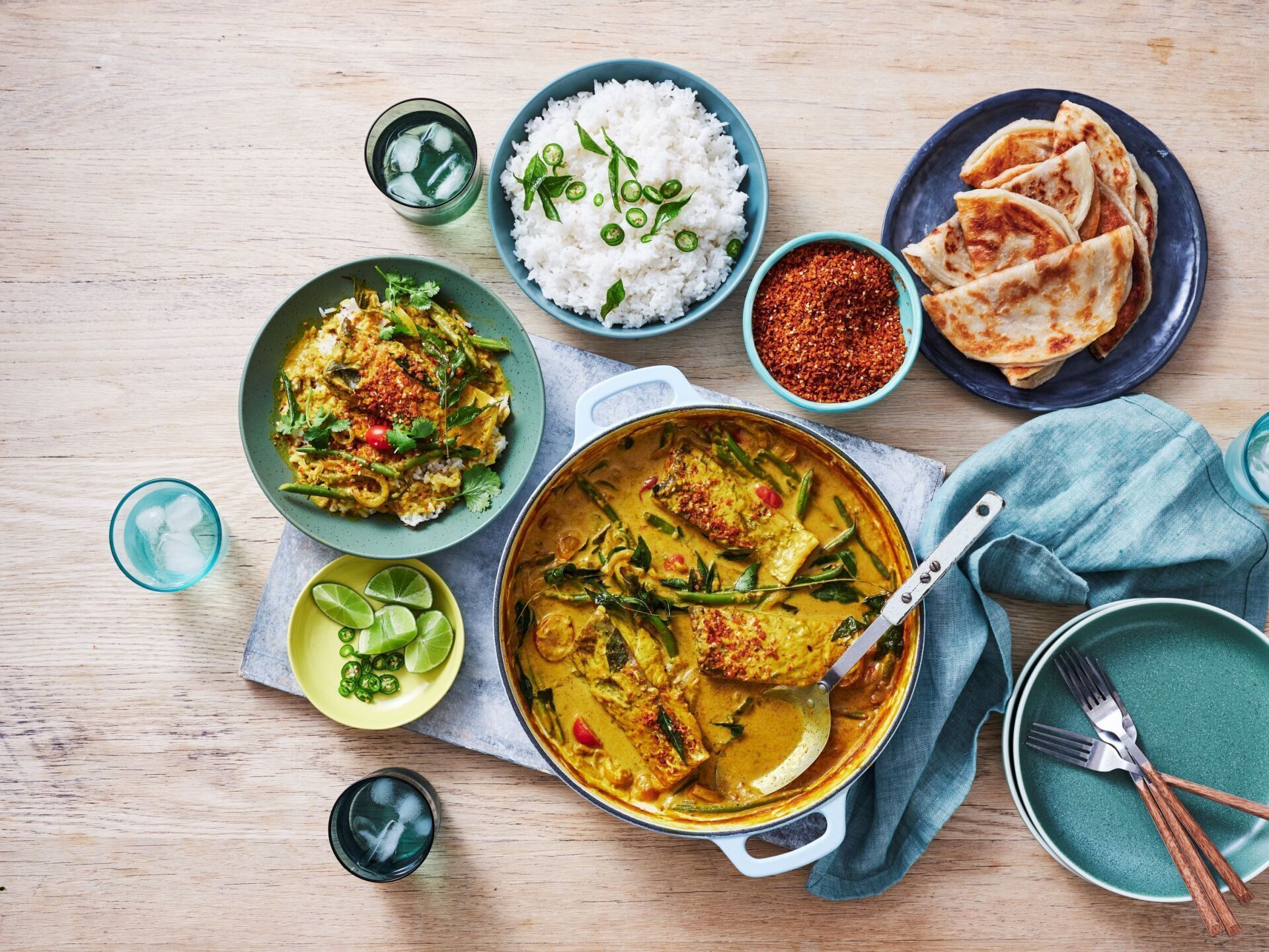 A meal of yellow curry with fish, rice, flatbread, and lime wedges arranged on a wooden table with plates and glasses.