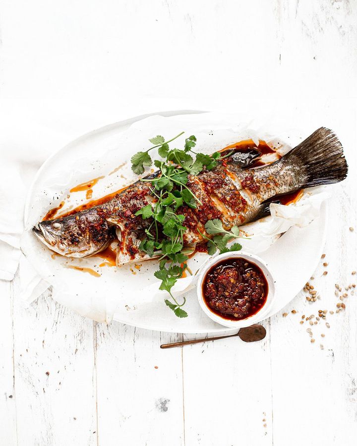 A whole cooked fish topped with cilantro and red sauce on a white platter, served with a small bowl of chili sauce.