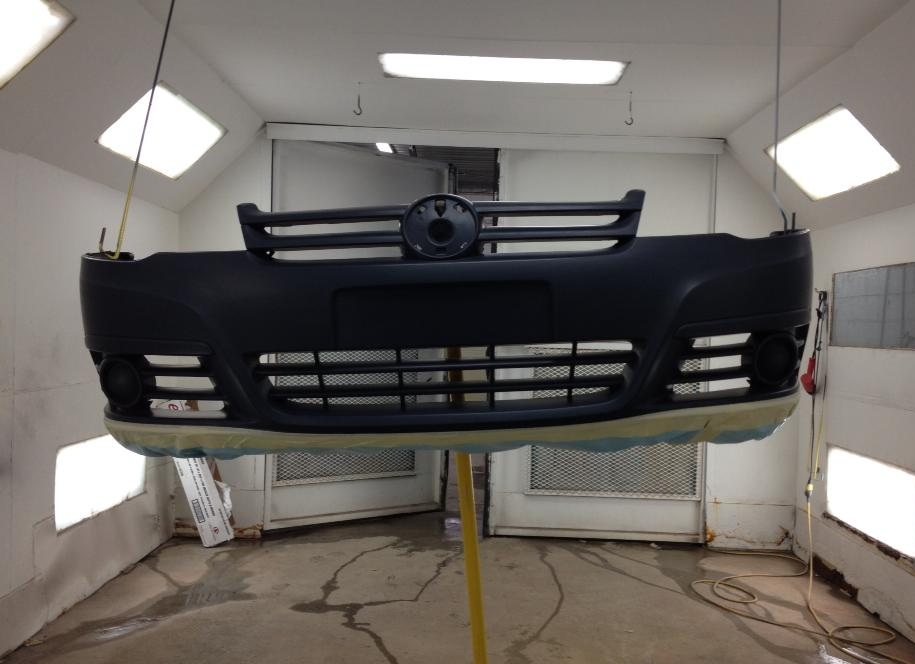 Pare-chocs et calandre de voiture noire suspendus dans une cabine de peinture, avec éclairage mural et au plafond.