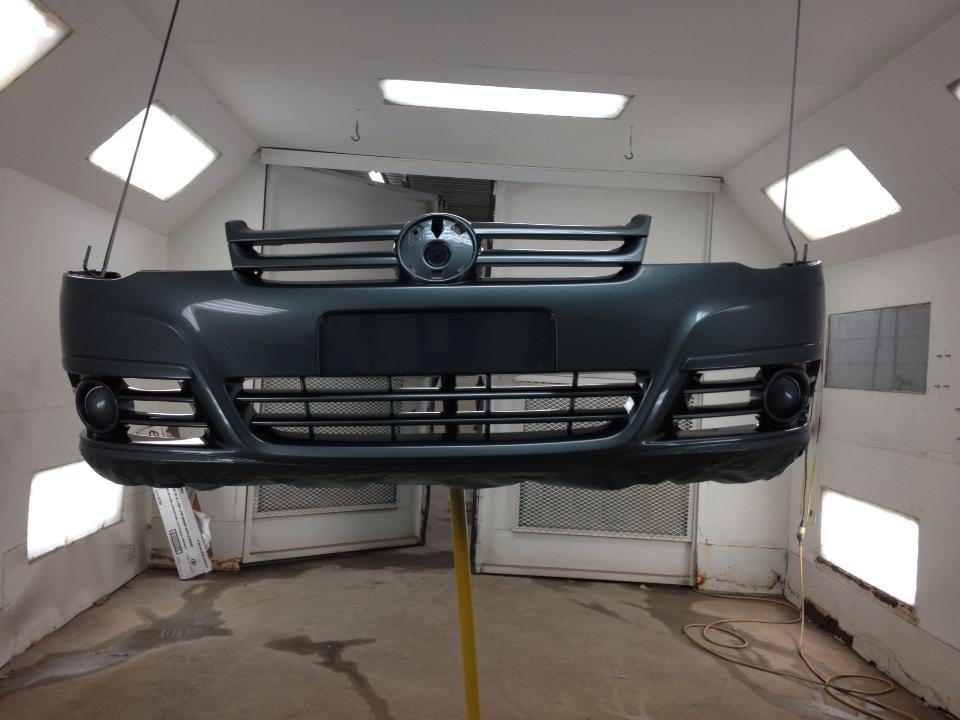 Pare-chocs avant noir d'une voiture suspendu dans une cabine de peinture automobile éclairée par des lumières vives et aux murs blancs.