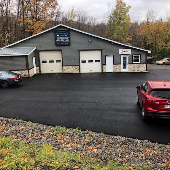 Atelier automobile gris avec deux portes de garage, des voitures garées et un parking asphalté mouillé en automne.