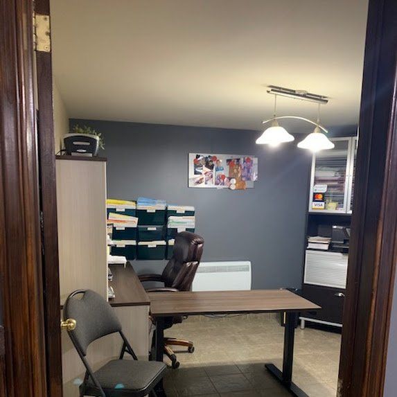 Petit bureau avec bureaux, chaises, étagères à dossiers et un mur gris, vu à travers une porte.