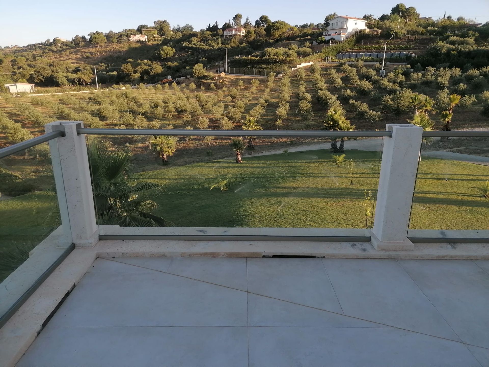 Vista dal balcone: ringhiera in vetro, campo verde, ulivi su una collina sotto un cielo limpido.