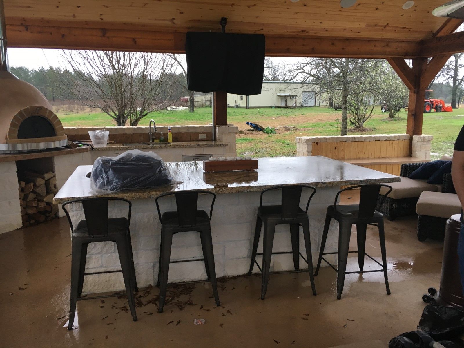 Outdoor kitchen with bar seating, pizza oven, and TV; covered patio overlooking a grassy area.