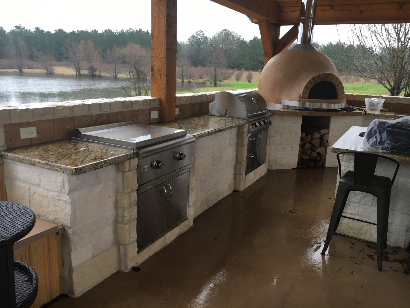 Outdoor kitchen with granite countertops, grill, oven, and wood-fired pizza oven overlooking a lake.