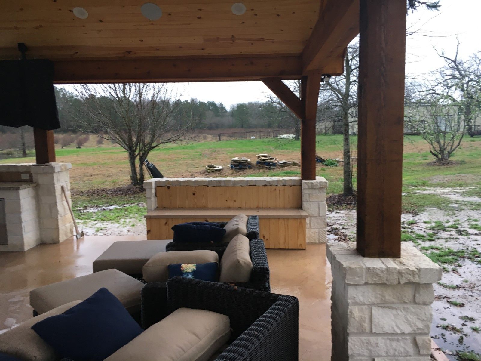 Covered outdoor patio with seating, wooden beams, stone columns, and view of a field.