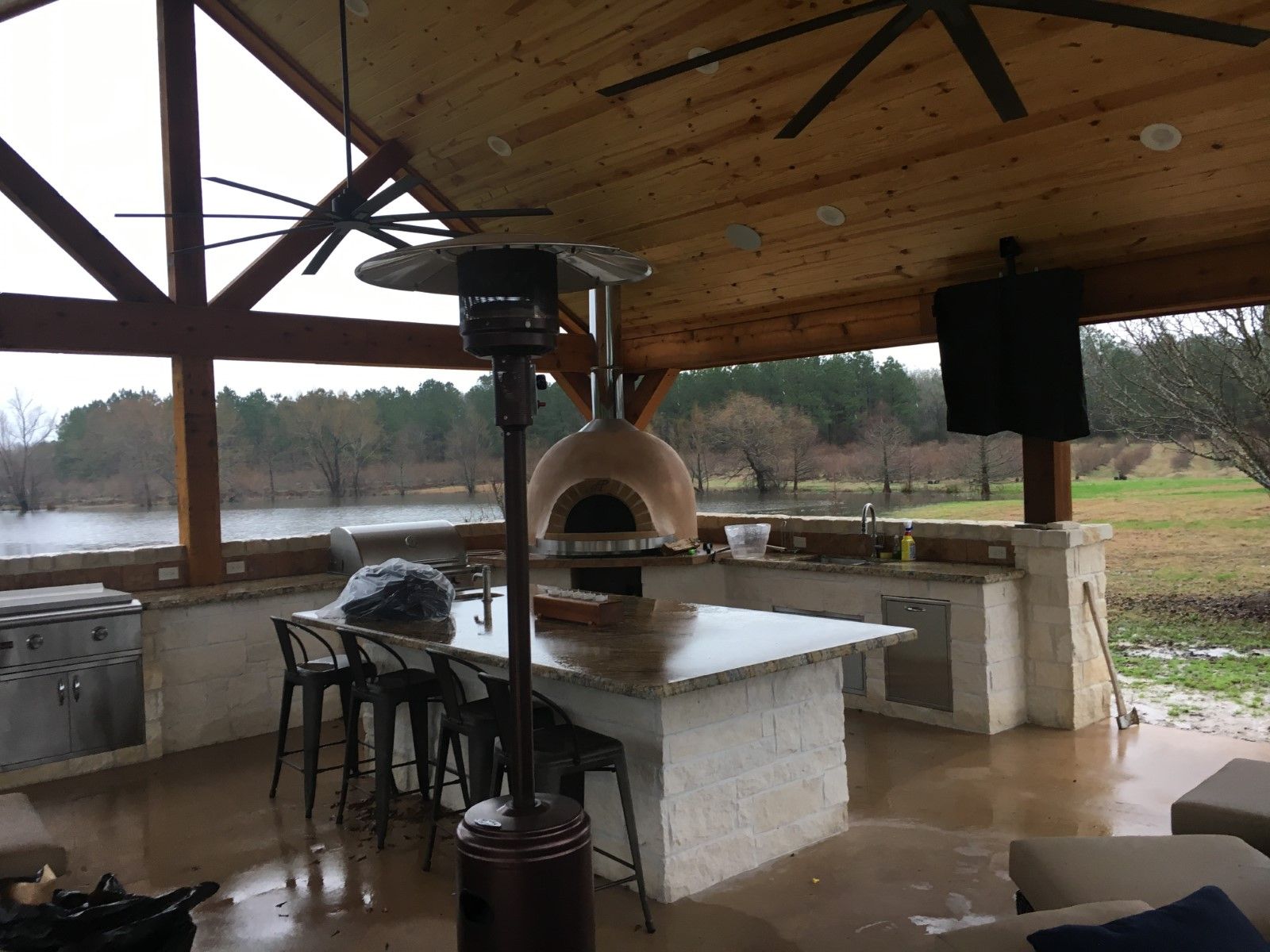 Outdoor kitchen under a wooden-beamed roof with an oven, bar, chairs, and a flooded landscape in the background.