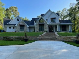 Elegant two-story house with stone and light gray exterior, featuring a long driveway, steps, and well-manicured lawn under a sunny sky.