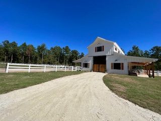 White barn with a covered porch, gravel driveway, and white fence under a blue sky.