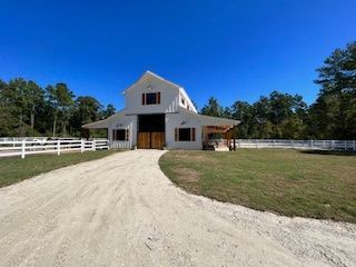 White barn with wooden doors, a gravel driveway, and a white fence under a blue sky.
