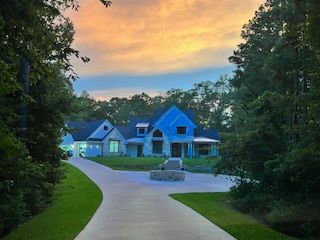 A large house with blue roof and stone facade, set at the end of a long driveway with trees and sunset sky.