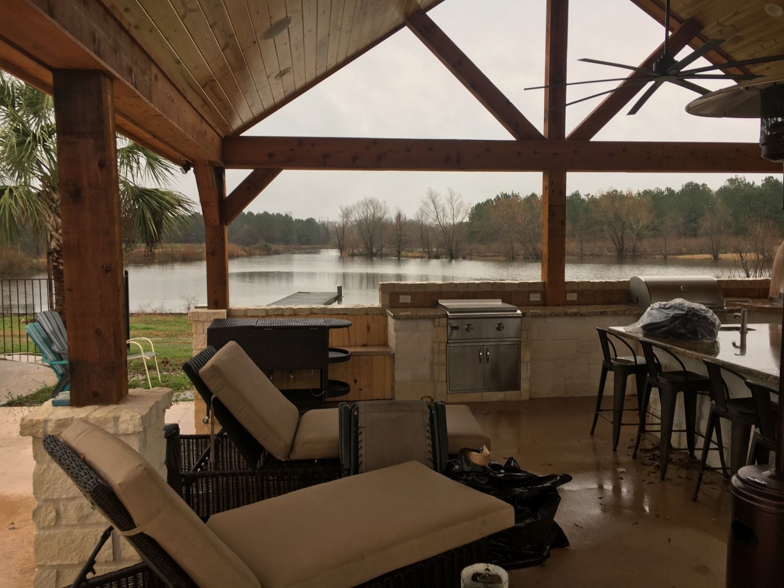 Covered patio overlooking a lake, with outdoor kitchen, seating, and exposed wooden beams.