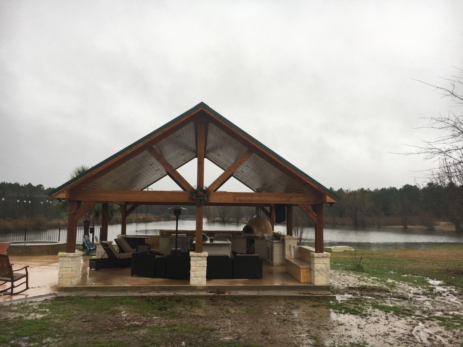 Outdoor pavilion with seating area, stone pillars, wooden frame, and lake in background under a cloudy sky.