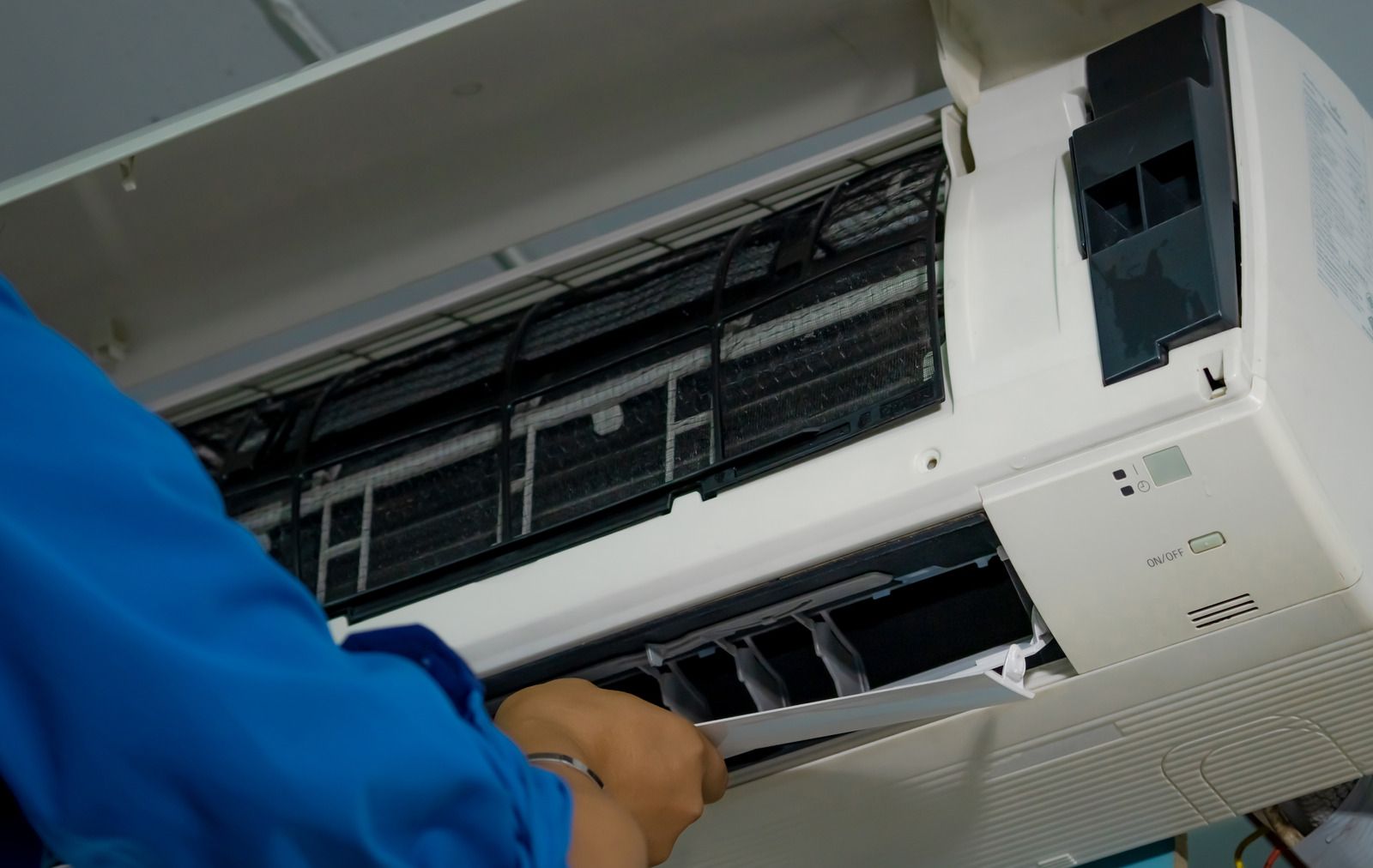 A person in a blue shirt performs maintenance on a white wall-mounted air conditioning unit.