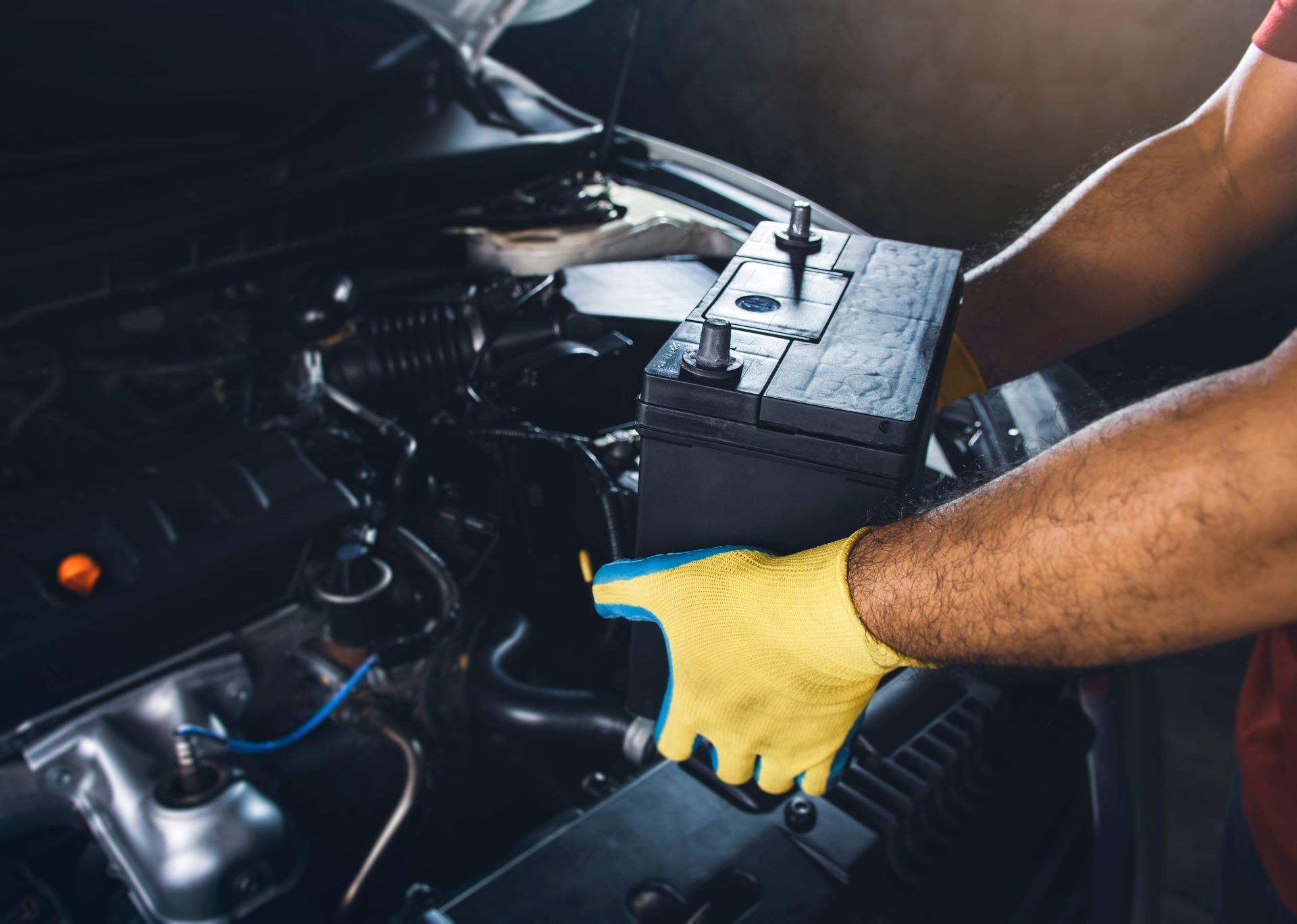 Mechanic's gloved hand holding a car battery beside an open vehicle engine bay