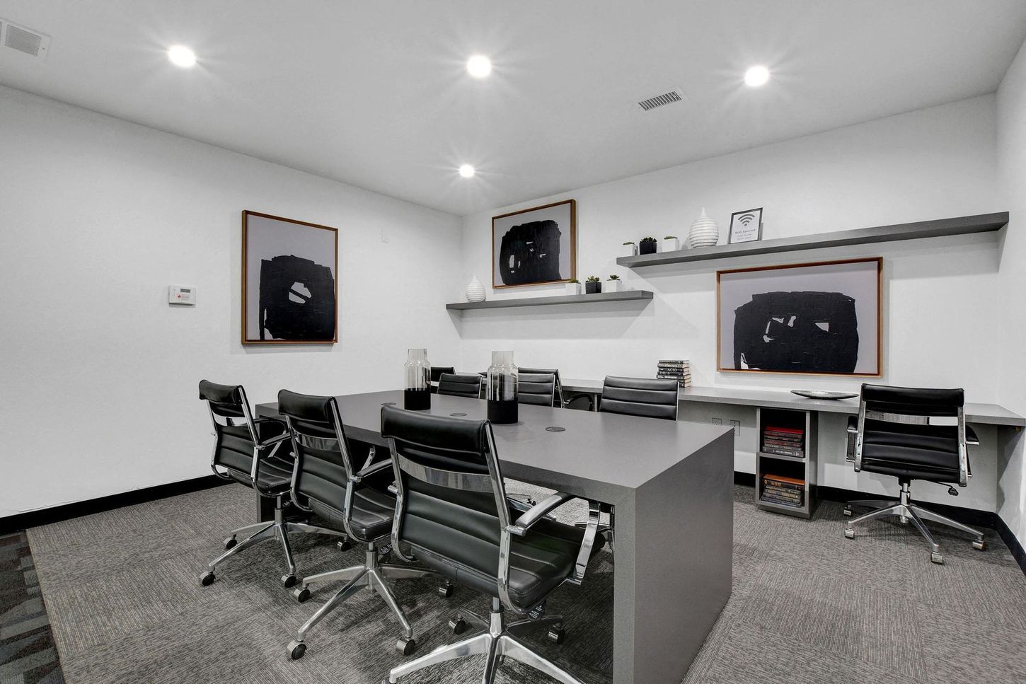 A modern office meeting room with gray table and chairs, artwork, shelves, and white walls.