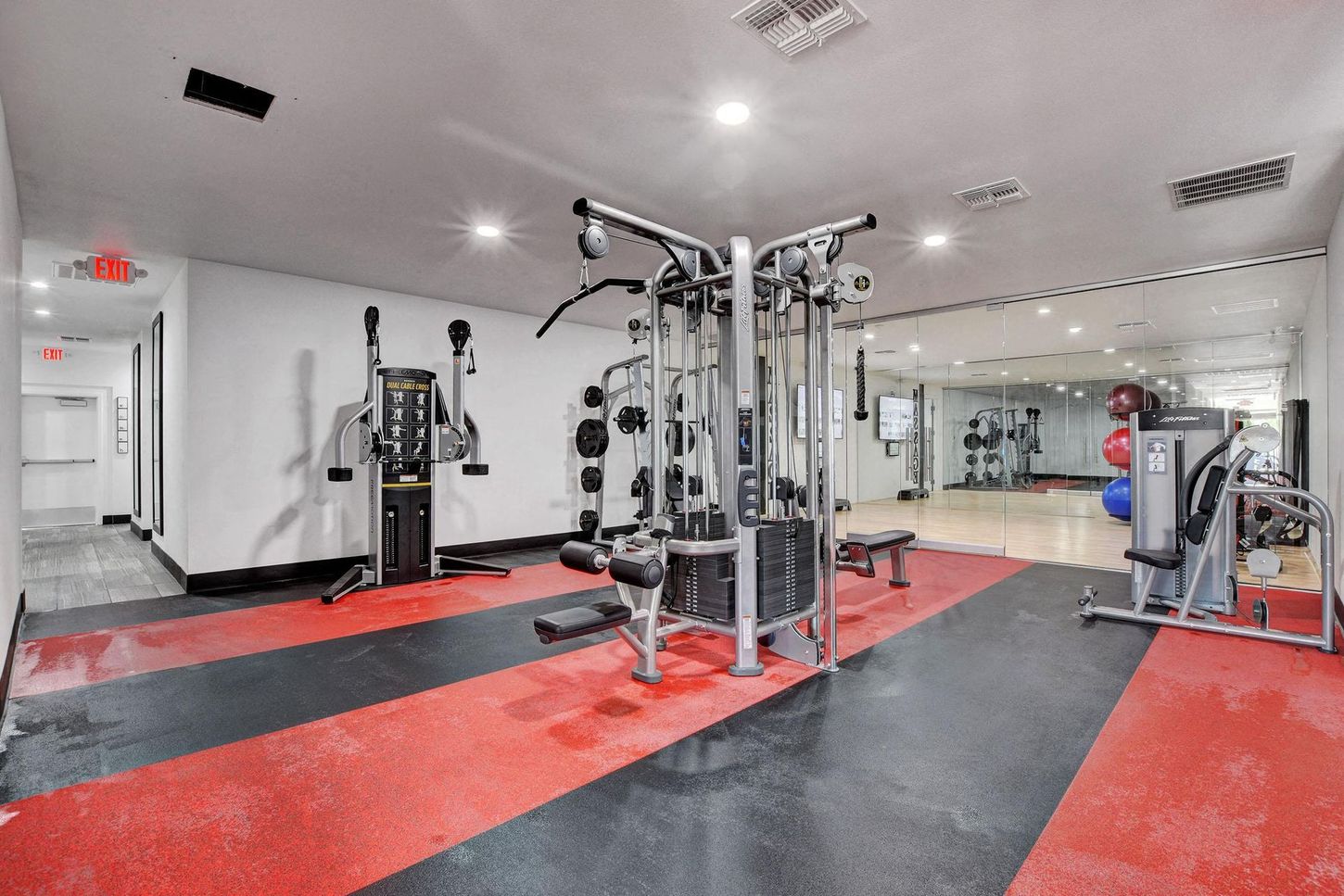 Gym interior with workout machines on red and black flooring, mirrored wall.