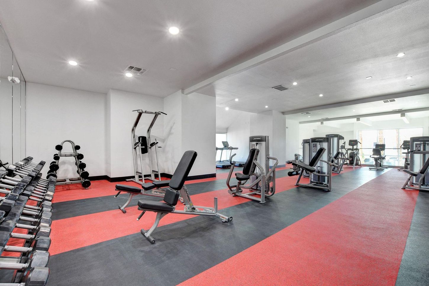 Gym interior with weight racks, weight machines, red and black flooring, and a mirrored wall.