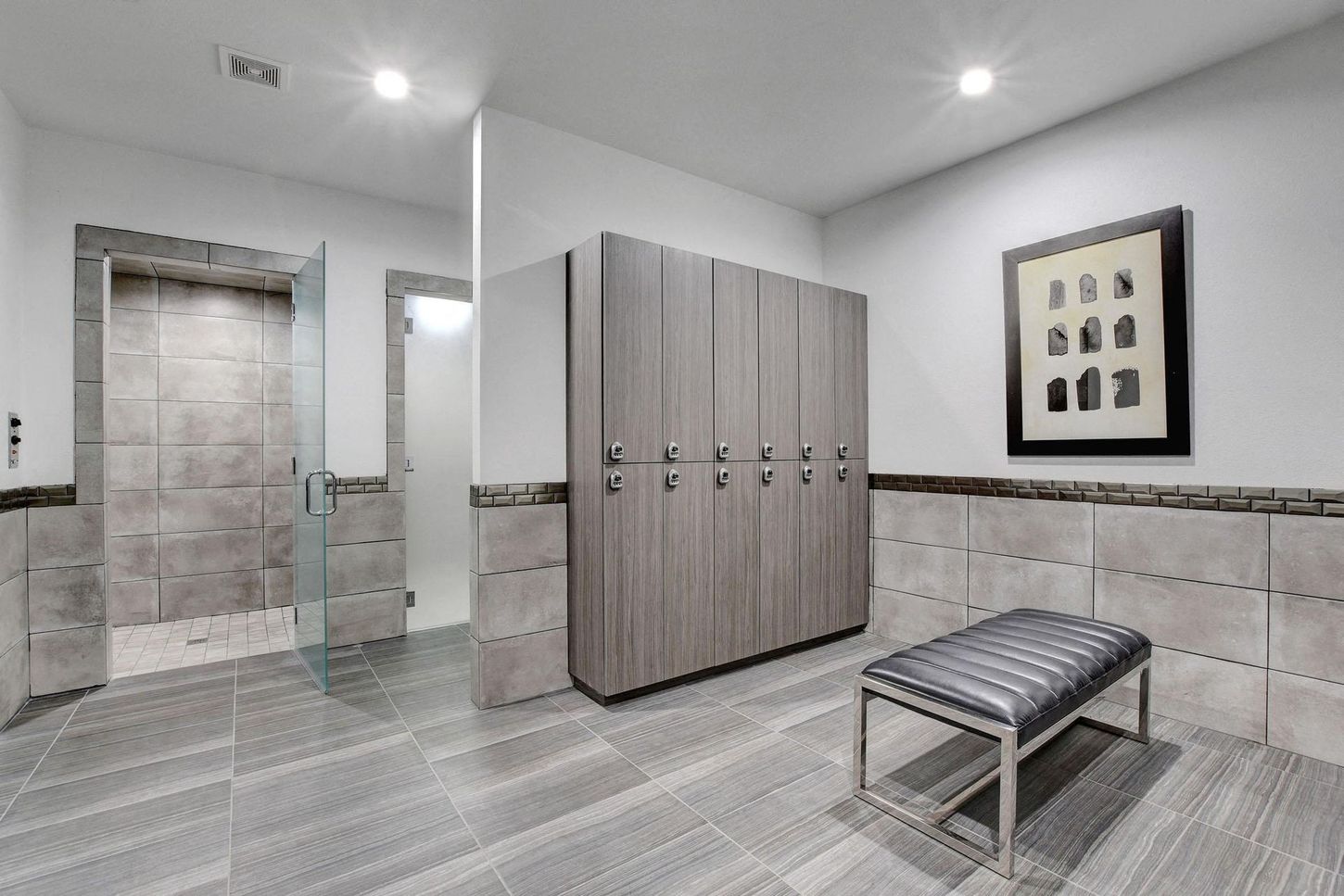 Locker room with gray lockers, bench, tiled floors, and a glass shower.
