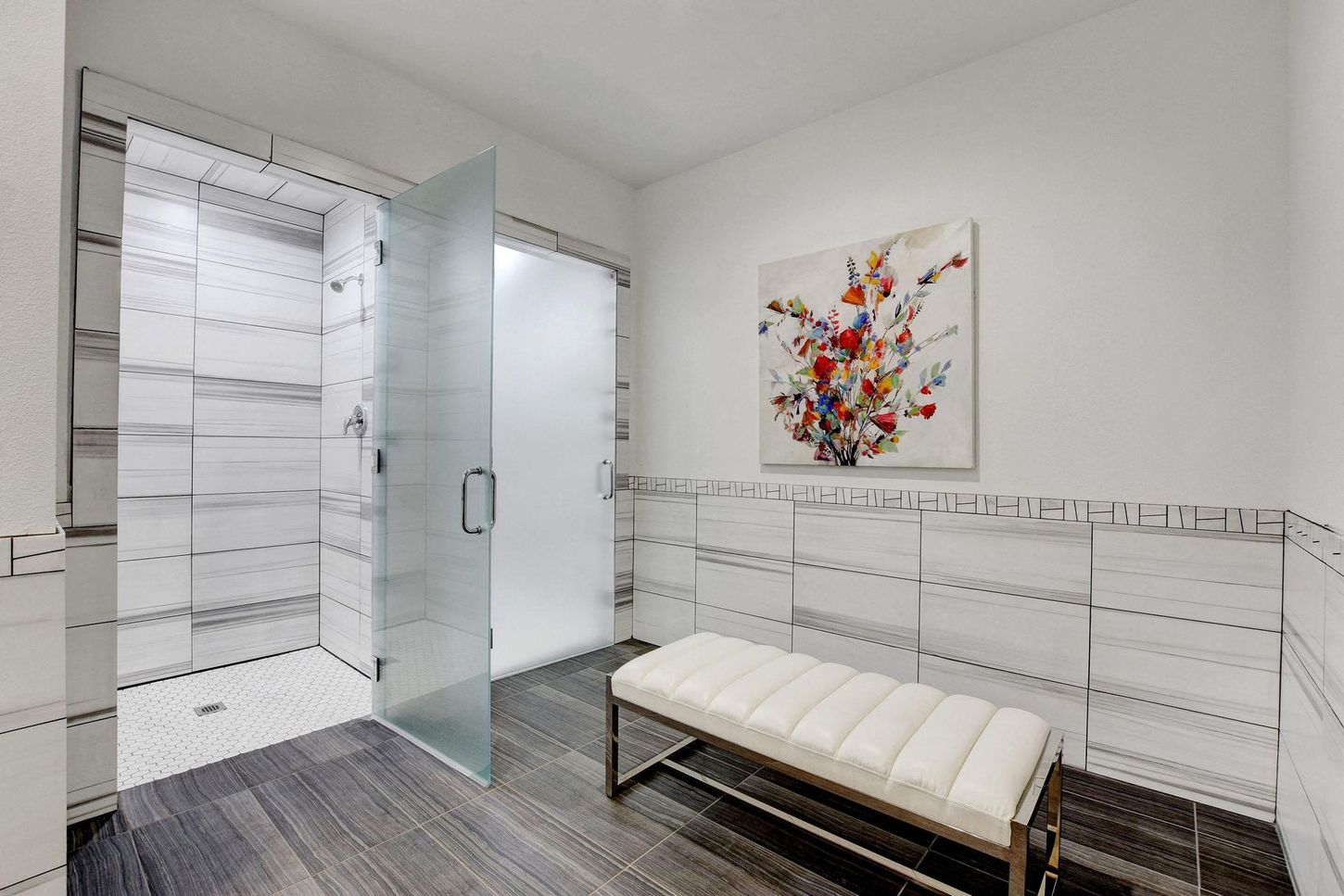Modern bathroom with a glass shower, bench, and artwork; white and gray tile with dark wood-look floor.