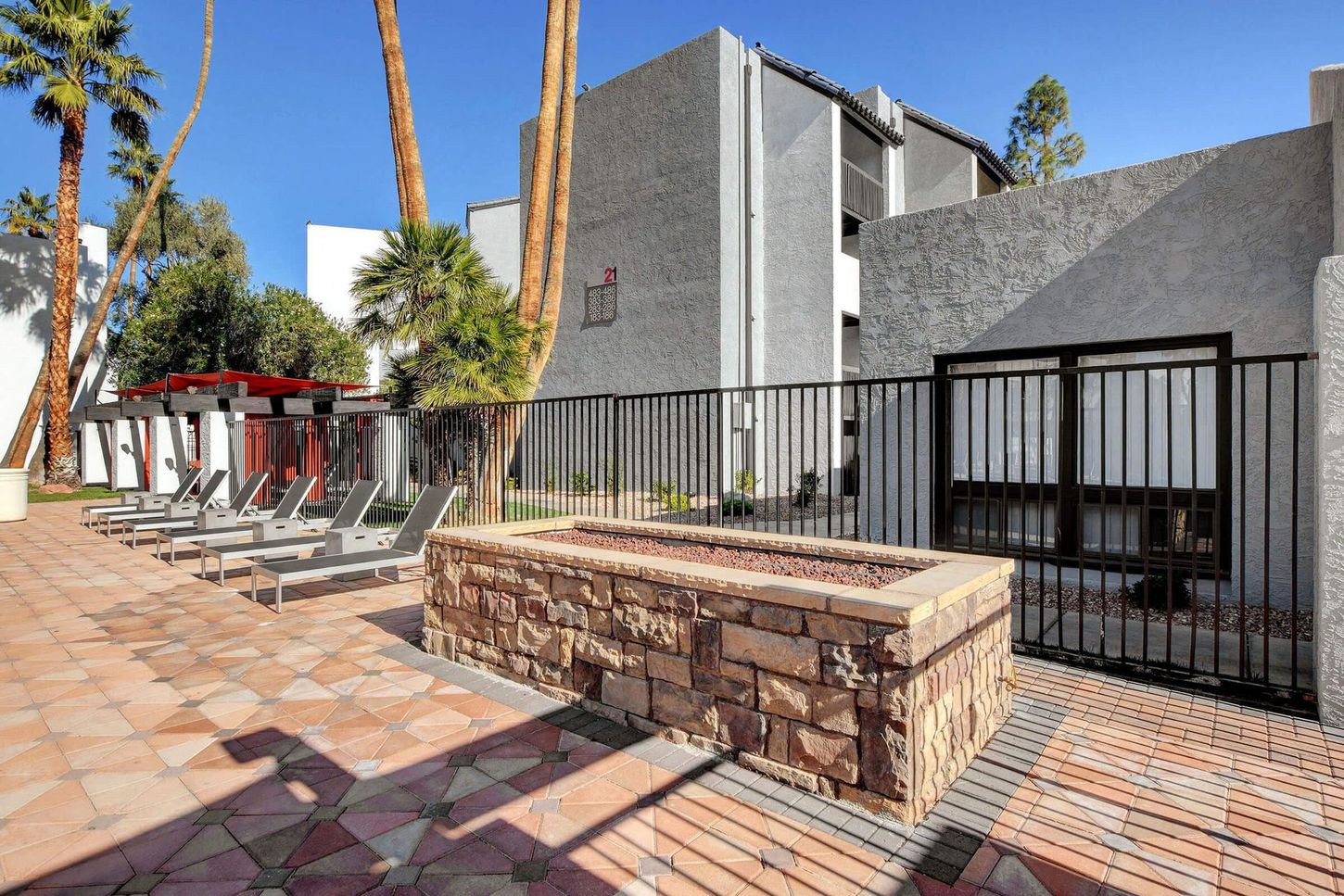 Outdoor area with fire pit, lounge chairs, and building in the background, under a blue sky.