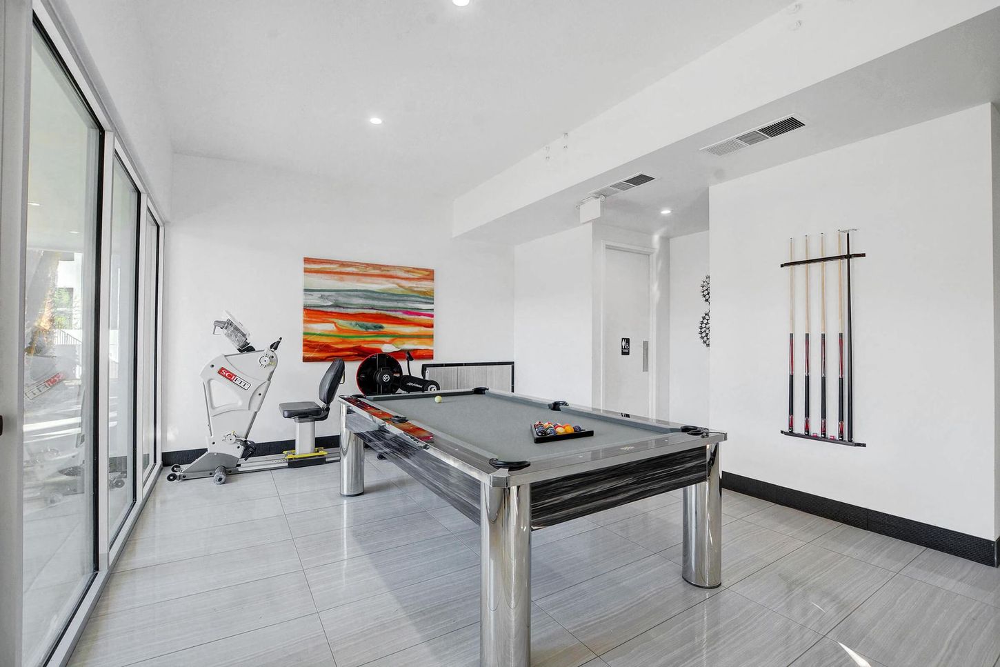 Pool table in a bright room with exercise equipment, art, and cue sticks on the wall.