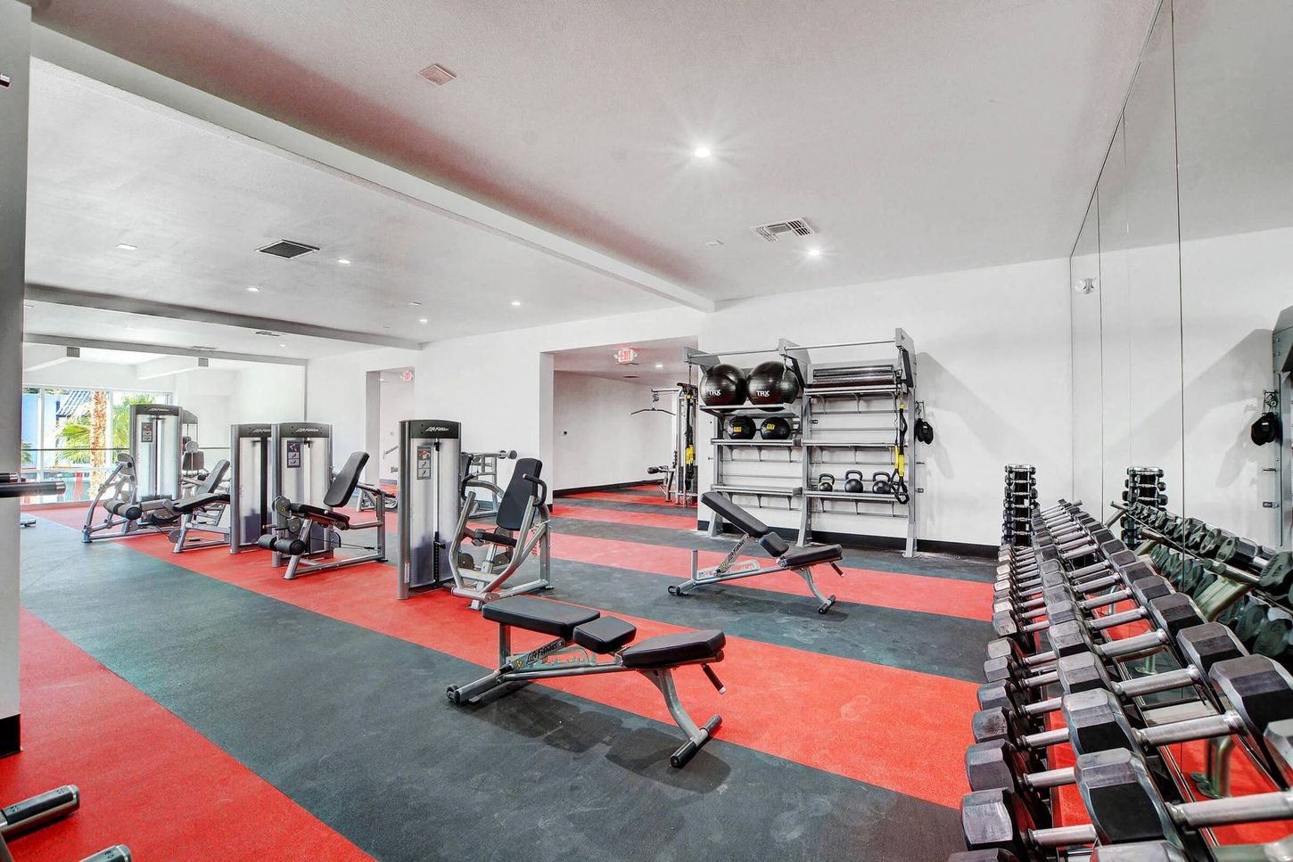 Gym interior with workout machines, weights, and red and black flooring.